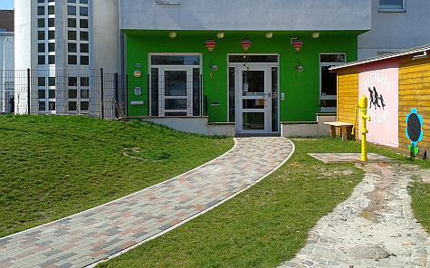 Grünes Gebäude zu dem ein gepflasterter Weg durch eine Rasenfläche führt. Rechts steht ein Holzschuppen.