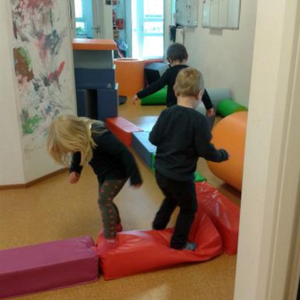 3 Kinder, die auf großen bunten Kissenbausteinen in einer Kita laufen und spielen.
