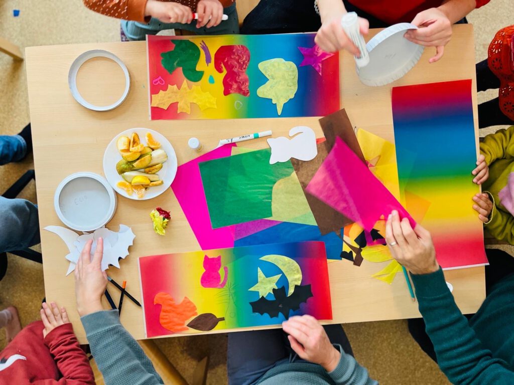 Draufsicht eines Tisches, auf dem buntes Bastelpapier liegt. von den Bildränder ragen Unterarme und Hände von Kindern sowie von einer erwachsenen Person mit Ring am Finger in das Bild.