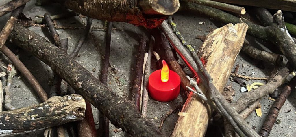 Auf grauem Boden liegen trockene, grau-schwarze und braune Äste. In der Mitte leuchtet ein rotes elektrisches Teelicht.