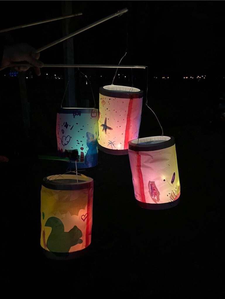 vor schwarzem Hintergrund leuchten vier selbstgebastelte Laternen in Rot-, Gelb- und Blautönen. Auf dem roten, gelben und blauen Transparentpapier der Laternen sind schemenhaft Figuren zu erkenne: ein Eichhörnchen, Vögel.