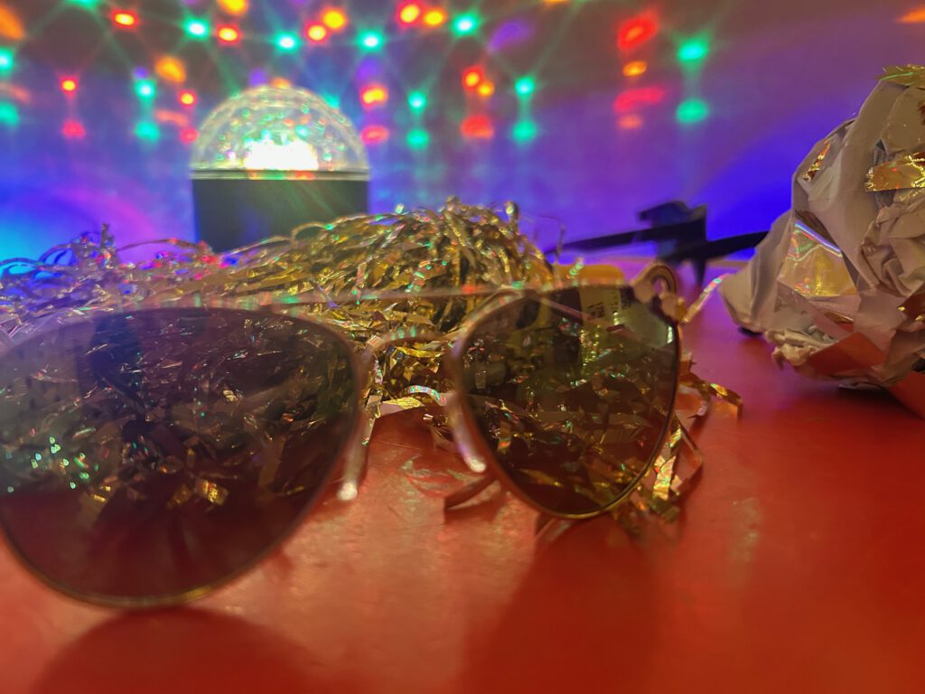 Lichtinstallation. Im Vordergrund eine Flieger-Sonnenbrille, dahinter ein goldener Pompom, im Hintergrund eine Diskokugel, die kreisförmig rote und grüne Lichtpunkte an die Wand dahinter wirft