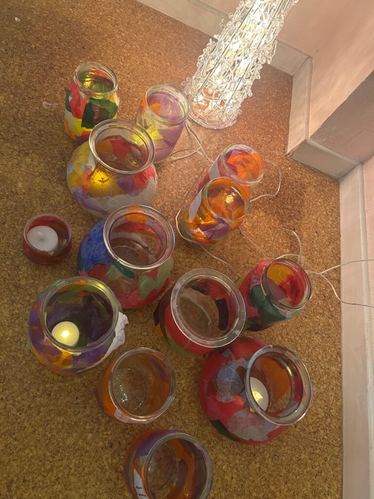 Draufsicht. Auf einem Korkfußboden stehen Gläser unterschiedlicher Formen und Größen, die von außen mit Transparentpapier beklebt wurden. In einigen der Gläser leuchten elektrische Teelichter. In der oberen rechten Bildecke leuchtet eine elektrishce Lichterpyramide.