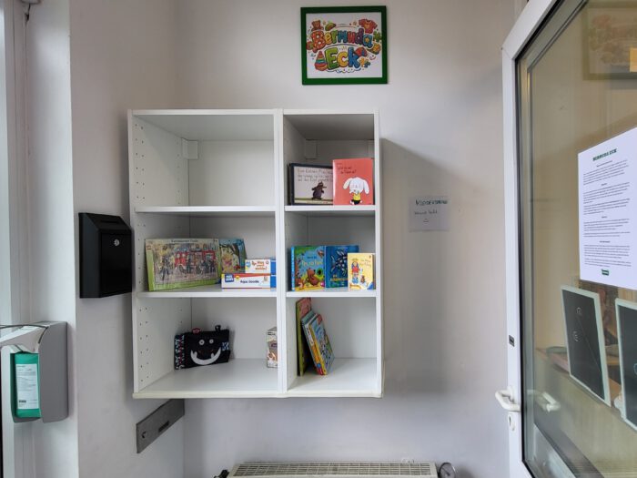 Ein weißes Regal an einer Wand mit einigen Büchern und Spielsachen. Darüber steht Bermuda Eck.