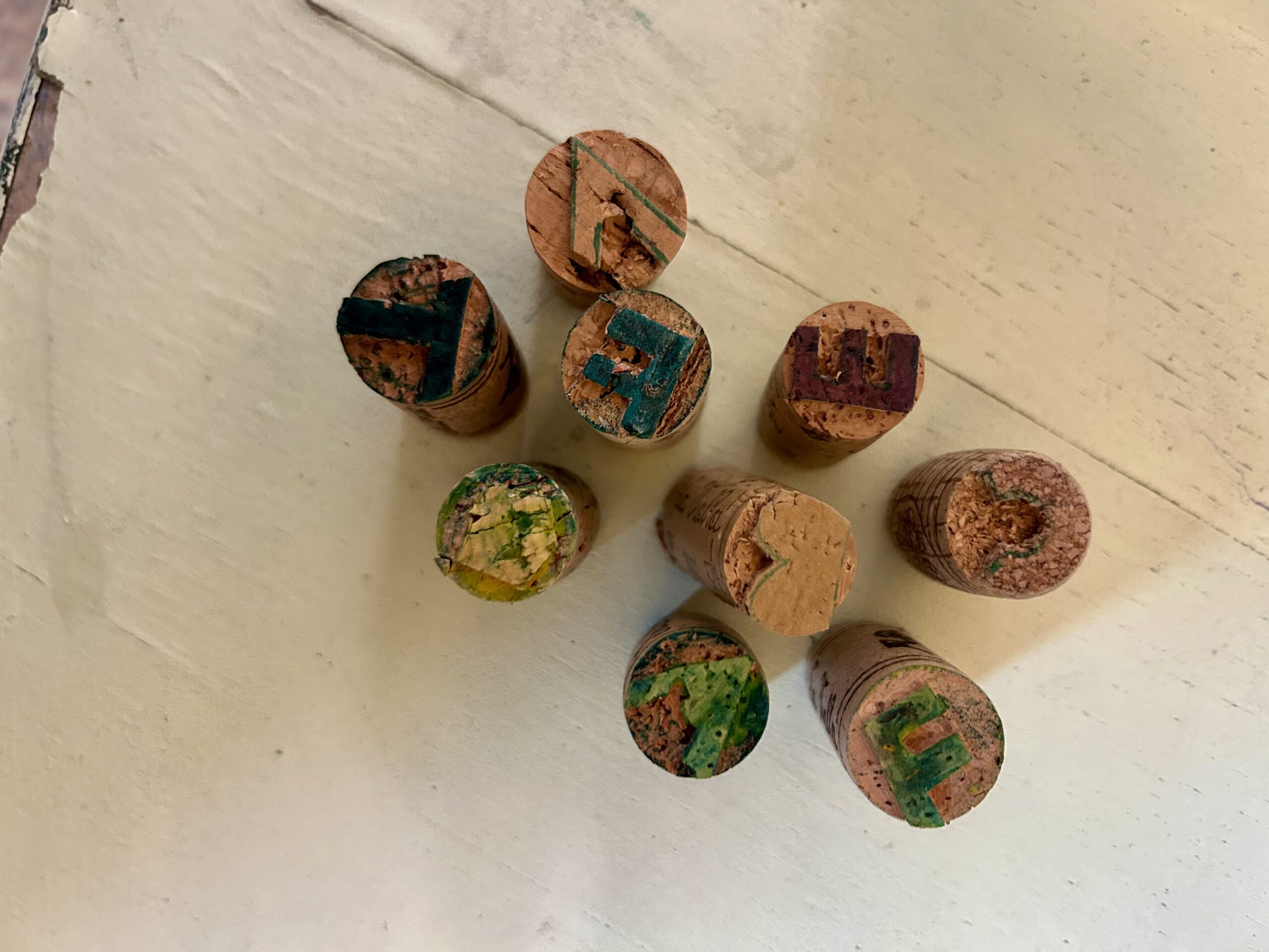 neun stehende Flaschenkorken, in deren obere Enden verschiedene Motive geschnitzt und mit Farbe benetzt sind