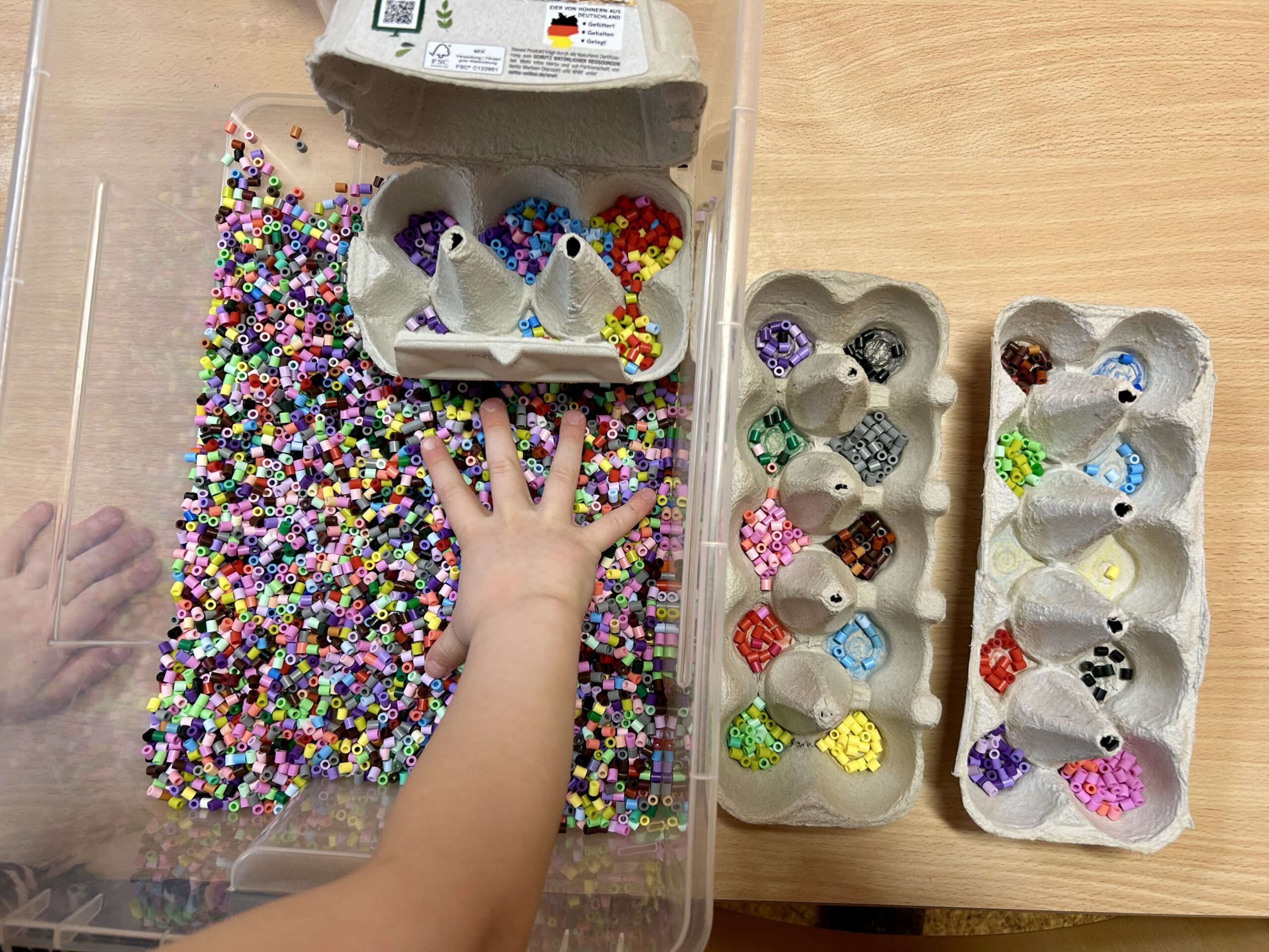 eine Kinderhand greift in eine durchsichtige Kunststoffkiste voller farblich unsortierter Bügelperlen. In der Kunststoffkiste steht ein 6er-Eierpappkarton, in dem Bügelperlen farblich sortiert sind. Neben der Kunststoffbox stehen zwei 10er-Eierpappkartons mit ebenfalls farblich sortierten Bügelperlen.